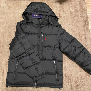 Svart dunjacka från Polo Ralph Lauren - Svart dunjacka från Polo Ralph Lauren med huva, dragkedja och flera fickor. Jackan har quiltad design och en röd Polo-logga på bröstet. Snygga detaljer som snörning vid huvan och RL-knappar. Perfekt för kalla dagar och riktigt stilren look. Med avtagbar luva. Tags medföljer. Fint skicka stl S passar bra på mig är ca 175. Justerbar midja. 