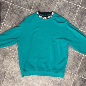 Turkos sweatshirt från Acne Studios - Säljer en chill turkos sweatshirt från Acne Studios med rund hals och coolt mönstrad krage i svart och vitt. Tröjan har långärmad passform och är gjord i mjuk bomull, perfekt för att dra på sig när man vill vara bekväm men ändå se snygg ut.