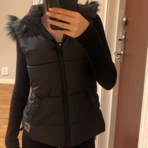 Svart väst med päls från H&M - Svart väst från H&M med avtagbar huva och fuskpälsdetaljer. Västen har  två fickor med knappar och quiltad design. Perfekt för lager-på-lager under höst och vinter. Snygg och praktisk.