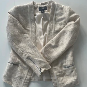 Beige kavaj med dragkedjor från H&M - Snygg beige kavaj från H&M i storlek 36. Jackan har ett vävt tyg med diskreta guldtrådar, silverfärgade dragkedjor på fickorna och är helfodrad. Modellen är öppen framtill och har långärmade ärmar. Perfekt för dig som gillar stilrena detaljer. Aldrig använd!