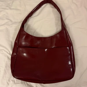 Mörkröd axelväska i skinn - Snygg mörkröd axelväska i glansigt skinnmaterial med stilren, rundad form. Väskan har ett stort huvudfack med ljusgrått innerfoder och en praktisk innerficka med dragkedja. Perfekt storlek för att få plats med allt du behöver och enkel att slänga över axeln.