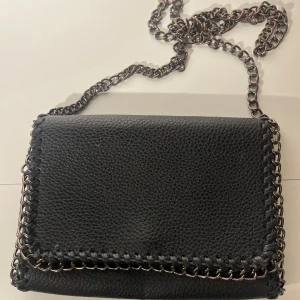 Svart axelväska med kedjedetaljer - Cool svart axelväska i skinn med grov kedja som axelrem och runt kanterna. Väskan har ett lock med magnetknäppning och insida med dragkedjefack. Perfekt storlek för det viktigaste och ger en edgy vibe till din outfit.