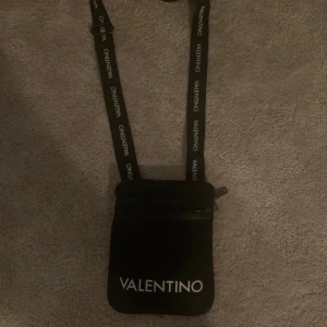 Svart axelväska från Valentino - Snygg svart axelväska från Valentino med justerbar rem där loggan är tryckt i vitt. Väskan har en praktisk dragkedja upptill och ett stilrent, minimalistiskt utseende. Perfekt storlek för mobil, plånbok och småsaker. Enkel att matcha med olika outfits.