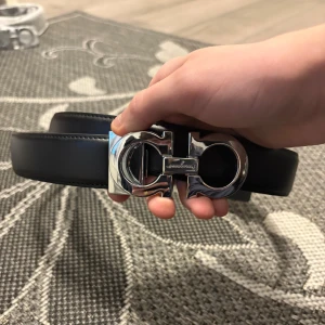 Svart skärp från Salvatore Ferragamo - Snyggt svart bälte från Salvatore Ferragamo med bred rem i slätt skinn och ikoniskt silverfärgat spänne med logga. Perfekt accessoar för att lyfta din outfit med en lyxig touch. Klassisk design som funkar till många stilar.