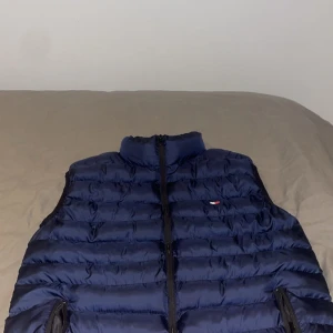 Mörkblå dunväst från Tommy Hilfiger - Snygg mörkblå dunväst från Tommy Hilfiger med quiltad design och hög krage. Västen har dragkedja framtill och två praktiska fickor med dragkedja. Liten Hilfiger-logga på bröstet ger en clean och sportig look.