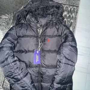 Svart pufferjacka från Polo Ralph Lauren - Svart pufferjacka från Polo Ralph Lauren med huva och dragkedja framtill. Jackan har ett rött Polo-logomärke på bröstet och två sidofickor med dragkedja. Materialet är syntet och jackan har en normal passform, perfekt för kalla dagar.