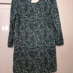 Klänning - Snygg klänning från Jacqueline de Yong i storlek 38. Klänningen har ett unikt abstrakt mönster i mörkblått, grönt och vitt, med långärmade ärmar och en liten v-ringning framtill. Tillverkad i mjuk polyester som känns skön mot huden.