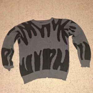 Grå och svart stickad tröja Manfinity - Säljer en grå stickad tröja från Manfinity i storlek M. Tröjan har ett unikt svart mönster som påminner om zebra eller abstrakta former. Rund halsringning och ribbade muddar vid ärmslut och nederkant. 
