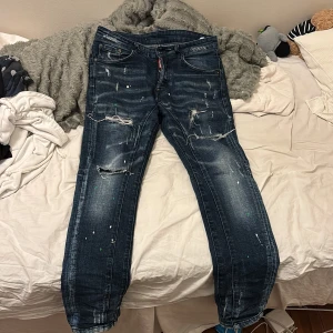 Dsquared2 blå distressed jeans - Säljer ett par blå jeans från Dsquared2 med coola slitningar, färgstänk och trasiga detaljer på benen. Jeansen har klassisk femficksdesign, normal passform och en snygg tvättad look. Perfekt för dig som gillar streetstyle och vill sticka ut.