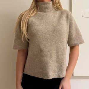 Beige stickad polotröja kort ärm - Supermysig beige stickad polotröja med korta ärmar och hög krage. Tröjan har en rak passform och ribbade kanter vid ärmslut, hals och nederkant. Perfekt för lager-på-lager under höst och vår.