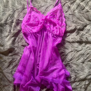 Lila spetstopp med rosett - Supersöt lila topp  med tunna axelband och transparent mesh. Överdelen har vacker spets och en liten rosett framtill. Toppen har volangdetaljer längst ner och är perfekt för dig som gillar en feminin och lekfull stil.