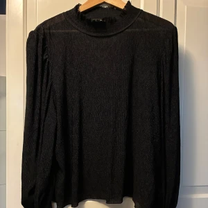 Svart glittrig blus med volang - Svart glittrig blus med långärmade puffärmar och smockade muddar med volangdetalj. Blusen har en hög krage med volangkant och ett tunt, skimrande tyg som ger en lyxig känsla.