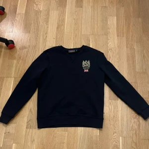 Svart sweatshirt från Morris, S - Svart sweatshirt från Morris med broderad guldfärgad logga och brittisk flagga på bröstet. Klassisk rund halsringning och ribbade muddar. Tillverkad i mjuk bomull för en skön känsla. Perfekt för dig som gillar stilrena och snygga plagg.