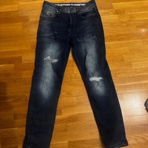 Mörkblå jeans från Hoodrich, storlek 30W - Snygga mörkblå jeans från Hoodrich med slitna detaljer på knäna och cool faded effekt. Klassisk femficksmodell med broderad logga på bakfickan och svart patch i midjan. Passformen är normal och materialet är robust denim. Perfekt för dig som gillar streetwear.