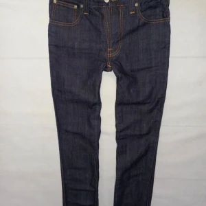 Nudie Jeans Slim Jim mörkblå jeans - Mörkblå jeans från Nudie Jeans, modell Slim Jim. Snygga kontrastsömmar i orange och klassisk femficksdesign. Bakfickorna har Nudie Jeans signaturbroderi. Tillverkade i 100% bomull med dry broken twill-väv. Slim passform för en modern look. Aldrig använt.