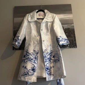 Vit och blå trenchcoat från Fontana 2.0 - Snygg vit trenchcoat från Fontana 2.0 med blå dekorativa mönster på ärmar och nederkant. Jackan har bred krage, knappar framtill och tillhörande bälte i midjan. Materialet är glansigt och känns lyxigt, perfekt för dig som vill sticka ut med stil.