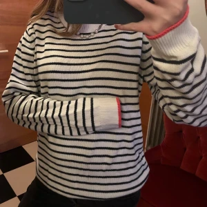 Randig vit stickad tröja med rosa kant - Snygg vit stickad tröja med svarta horisontella ränder och en poppande rosa kant vid ärmsluten. Tröjan har en klassisk rund hals och långa ärmar, perfekt för dig som gillar en chill och stilren look.