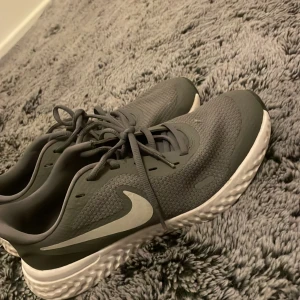 Grå Nike sneakers med vit sula - Snygga grå Nike sneakers med vit sula och klassisk swoosh på sidan. Skorna har snörning och är tillverkade i ett ventilerande syntetmaterial med stickad känsla. Perfekta för dig som gillar sportig stil och vill ha bekväma skor till vardagen.