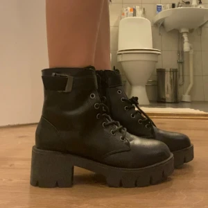 Svarta chunky boots med snörning - Säljer ett par svarta boots med grov sula och snörning framtill. Skorna har rund tå, klack och ett spänne vid ankeln som ger en cool vibe. Tillverkade i skinnimitation och passar perfekt till höstens outfits.