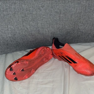 Röda Adidas F50 fotbollsskor - Säljer ett par röda Adidas F50 fotbollsskor med svarta detaljer och klassiska tre ränder på sidan. Skorna har en strömlinjeformad design utan snörning och är tillverkade i syntetmaterial för lätthet och snabbhet på planen. Perfekt för dig som vill sticka ut på fotbollsplanen.