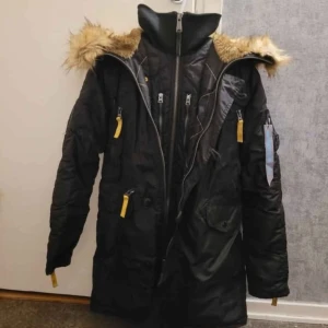 Svart parkas med päls och gula detaljer, Alpha Industries - Säljer en svart parkas från Alpha Industries med snygg fuskpäls på huvan och flera gula dragkedjor. Jackan har hög krage, många fickor och är perfekt för kalla dagar. Materialet är slitstark polyester och jackan är långärmad med normal passform.