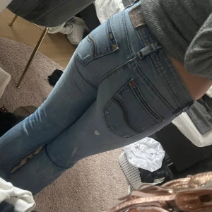 Ljusblå bootcut jeans från Lee - Säljer ett par ljusblå jeans från Lee med klassisk femficksdesign och låg midja. Jeansen har bootcut-ben och snygga slitningar framtill för en avslappnad vibe. Tillverkade i mjukt denimtyg som sitter skönt hela dagen.