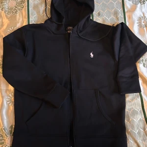 Svart hoodie från Polo Ralph Lauren - Svart hoodie från Polo Ralph Lauren med klassisk vit logga broderad på bröstet. Tröjan har huva, dragkedja framtill och två stora fickor. Perfekt för chill dagar och enkel att matcha med jeans eller joggers.
