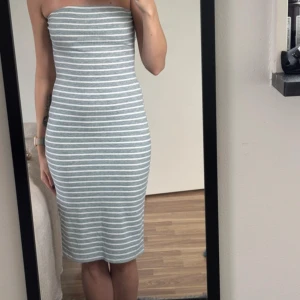 Randig axelbandslös klänning - Säljer en snygg axelbandslös klänning med horisontella vita och grå ränder. Klänningen är figurnära och har en midilängd som slutar strax under knäna. Materialet är ribbat och stretchigt, vilket ger en bekväm och tight passform.