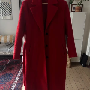 Röd kappa från & Other Stories - Säljer en snygg röd kappa från & Other Stories Paris Atelier. Kappan har klassisk krage, knappar framtill och ett matchande bälte i midjan. Långärmad och med fickor på sidorna. Materialet är mjukt och har en ullig känsla, perfekt för kyliga dagar.