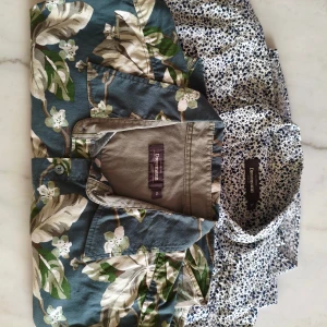 Två mönstrade skjortor Dressmann XL - Två kortärmade  skjortor från Dressmann i storlek XL. Ena skjortan har ett grönt och beige blommigt mönster på blå botten, den andra är vit med små svarta blommor. Båda har klassisk krage och knäppning framtill. Perfekta för dig som gillar mönster och vill sticka ut.