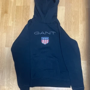 Svart hoodie från GANT - Svart hoodie från GANT med stort tryck på bröstet och ett broderat emblem i rött, vitt och blått. Tröjan har huva, magficka och långa ärmar. Perfekt för dig som gillar klassisk streetstyle och vill ha en bekväm och snygg look.