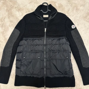 Moncler Cardigan - Svart Cardigan från Moncler med stickade ärmar och överdel, samt quiltad front i syntetmaterial. Jackan har två stora fickor med knappar, hög krage och hel dragkedja. Moncler-logga på vänster ärm. Perfekt för dig som gillar stilren och sportig look.