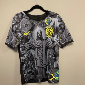 Brasil Nike fotbollströja med tryck - Unik Brasil fotbollströja från Nike med detaljerat svart och grått tryck av Kristusstatyn och brasilianska motiv. Gult Brasil-emblem och Nike-logga på bröstet, samt brasiliansk flagga på nederdelen. Kortärmad och tillverkad i lätt, ventilerande material.