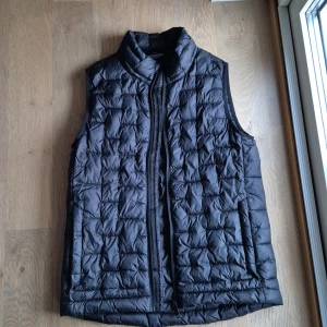 Svart quiltad dunväst från Everest - Svart dunväst från Everest med quiltad design och hög krage. Västen har dragkedja framtill och är tillverkad i syntetmaterial. Perfekt för lager-på-lager och har en clean, sportig look som funkar till många stilar.