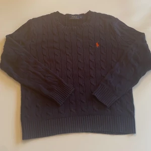 Mörkblå kabelstickad tröja Polo Ralph Lauren - Kabelstickad mörkblå tröja från Polo Ralph Lauren med rund halsringning och klassisk röd broderad logga på bröstet. Defekten ser ni på bild 4. Skriv om ni har funderingar!