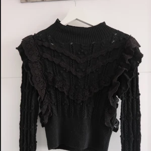 Svart stickad tröja med volang från Zara - Supersnygg svart stickad tröja från Zara med höghalsad krage, långa ärmar och detaljerade virkade volanger framtill och på axlarna. Tröjan har ett coolt strukturmönster och känns både trendig och unik. Perfekt för dig som gillar statement-plagg!