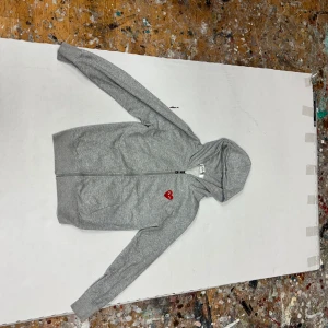 Grå hoodie från Comme des Garçons Play - Grå hoodie med dragkedja från Comme des Garçons Play. Tröjan har huva, långa ärmar och ett rött hjärta med ögon broderat på bröstet. Mjuk bomullskvalitet och ribbade muddar vid ärmslut och nederkant. Perfekt för chill dagar.