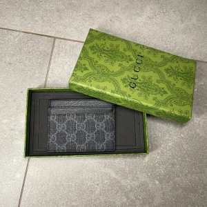 Svart plånbok  - Snygg svart plånbok med klassiskt mönster i grått. Slimmad design som passar perfekt för kort och sedlar. Kommer i original grön box med elegant mönster. Perfekt accessoar för dig som gillar lyxiga detaljer.