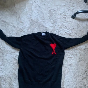 Svart stickad tröja från Ami - Svart stickad tröja från Ami med ett stort rött hjärta och bokstaven A broderat på bröstet. Tröjan har rund hals, ribbade muddar och lång ärm. Perfekt för dig som gillar stilrena plagg med en twist. Snygg att matcha med jeans eller chinos.