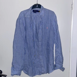 Randig blå skjorta från Ralph Lauren - Snygg blå och vit randig skjorta från Polo Ralph Lauren med klassisk krage och knappar framtill. Långärmad modell i lätt och skönt linnematerial, perfekt för varmare dagar. Diskret rosa logga broderad på bröstet ger en stilren touch.