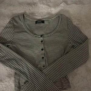 Randig långärmad topp från Bershka - Säljer en svartvit randig långärmad topp från Bershka med rund halsringning och knappar framtill. Toppen är ribbad och har en slim passform som sitter snyggt på kroppen. Perfekt för dig som gillar enkel och stilren design.