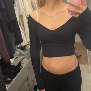 Svart croppad offshoulder topp - Säljer en svart croppad topp med offshoulder-design och långa ärmar. Toppen har snygg rynkning framtill och en djup v-ringning. Materialet är ribbat och stretchigt, vilket gör den bekväm och följsam. Perfekt för dig som gillar trendiga och stilrena plagg.