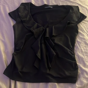 Svart blus med volang från Stradivarius - Snygg svart blus från Stradivarius i storlek M. Blusen har korta ärmar, en djup v-ringning med volangkant och en dekorativ knytning framtill. Materialet är mjukt och har en lätt glansig finish som ger en elegant vibe.