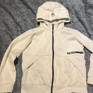 Beige hoodie från Sail Racing - Snygg beige hoodie från Sail Racing med svart dragkedja och logga på bröstet. Utsidan är slät medan insidan är mjukt fodrad i fleece. Tröjan har huva och långa ärmar, perfekt för kyliga dagar. Stilren design med praktiska fickor framtill.