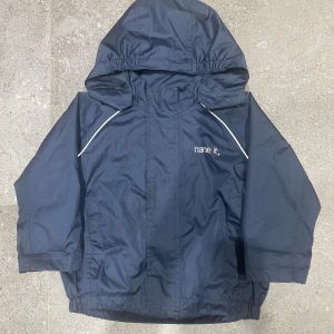 Mörkblå vindjacka från Name It - Snygg mörkblå vindjacka från Name It med huva och vita detaljer längs axlarna. Jackan har tryckta pilar på ryggen och elastisk nederkant. Perfekt för blåsiga dagar och enkel att matcha med jeans eller mjukis. Materialet är slitstark polyester. Storlek 92 för barn! 