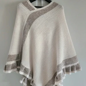 Beige stickad poncho med volangkant - Supermysig stickad poncho i beige och vitt med glittriga inslag i guld. Ponchon har breda ränder, volangkant nertill och en snygg omlottkänsla. Perfekt att slänga över en outfit för extra värme och stil.