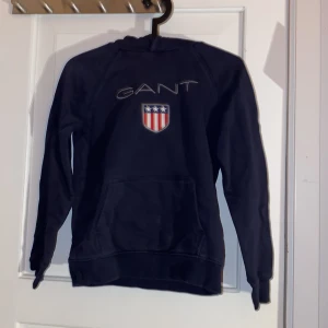 Mörkblå hoodie från GANT med logga - Säljer en mörkblå hoodie från GANT med broderad logga och amerikanskt inspirerat märke på bröstet. Tröjan har huva, magficka och långa ärmar. Perfekt för dig som gillar klassisk och sportig stil.