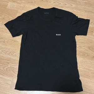 Svart t-shirt från BOSS - Säljer en stilren svart t-shirt från BOSS med diskret logga på bröstet. T-shirten har klassisk rund hals och korta ärmar, perfekt för en clean och enkel look. Materialet känns mjukt och bekvämt mot huden.