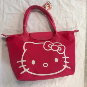 Hello Kitty rosa longchamp - Superfin rosa handväska från Hello Kitty med stort vitt tryck av Hello Kittys ansikte på framsidan. Väskan har två handtag, dragkedja och knappstängning upptill samt detaljer i syntetmaterial. Perfekt för dig som älskar kawaii och vill sticka ut. Mått ca: 40x25x14cm