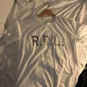 Ralph Lauren T-shirt - Vit t-shirt från RRL med tryck på bröstet. Klassisk rund hals och korta ärmar. Materialet känns mjukt och skönt, perfekt för en chill look. Trycket har en cool, lite sliten effekt som ger extra karaktär.